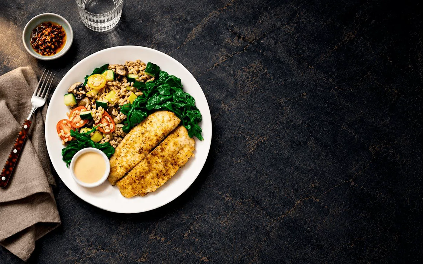 A CookUnity meal of crusted fish with farro, roasted vegetables and spinach on a dark table.